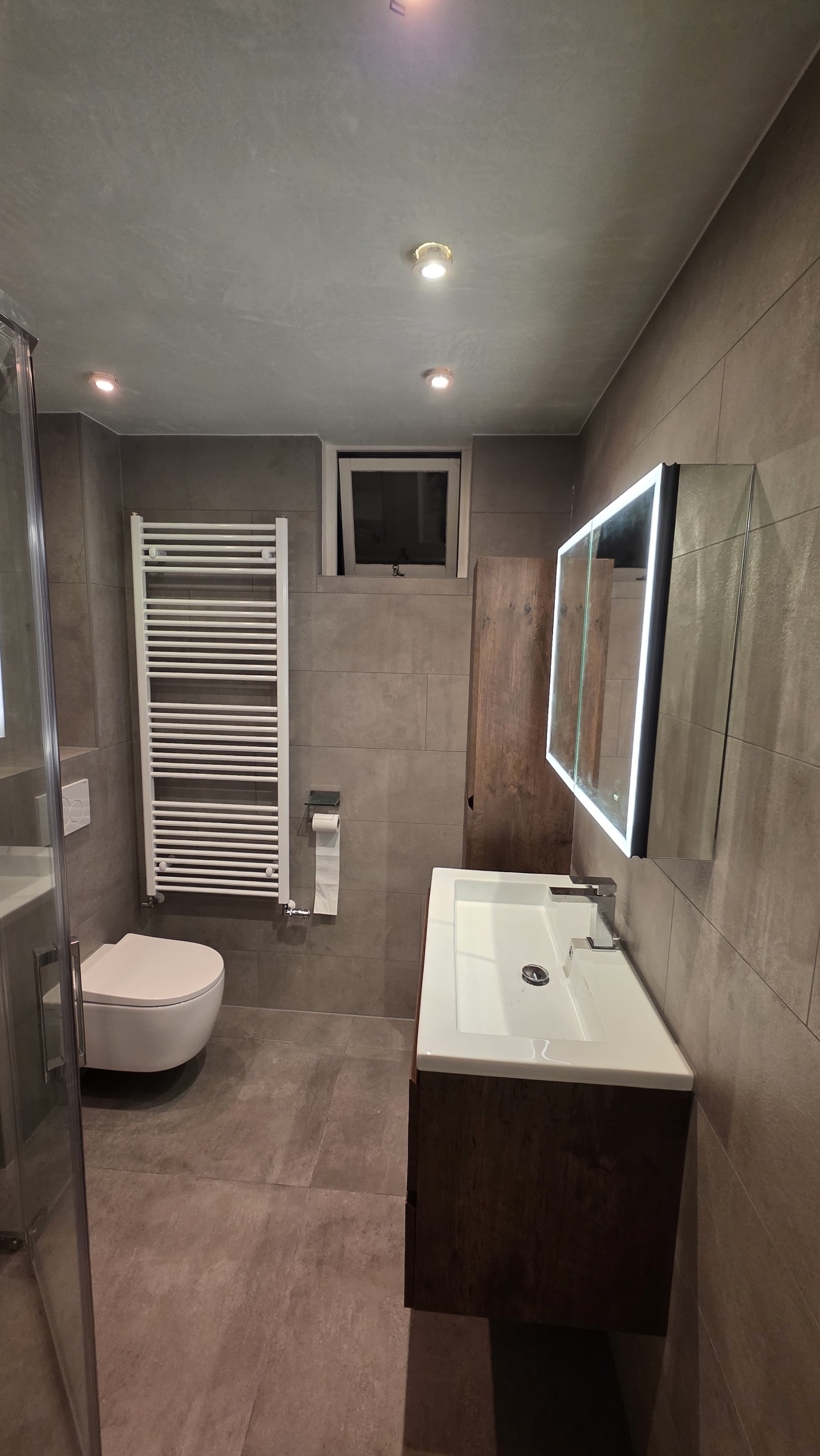 Afgewerkte badkamer met betonlook tegels, hangend toilet, handdoekradiator en notenhouten wastafelmeubel met ledspiegel