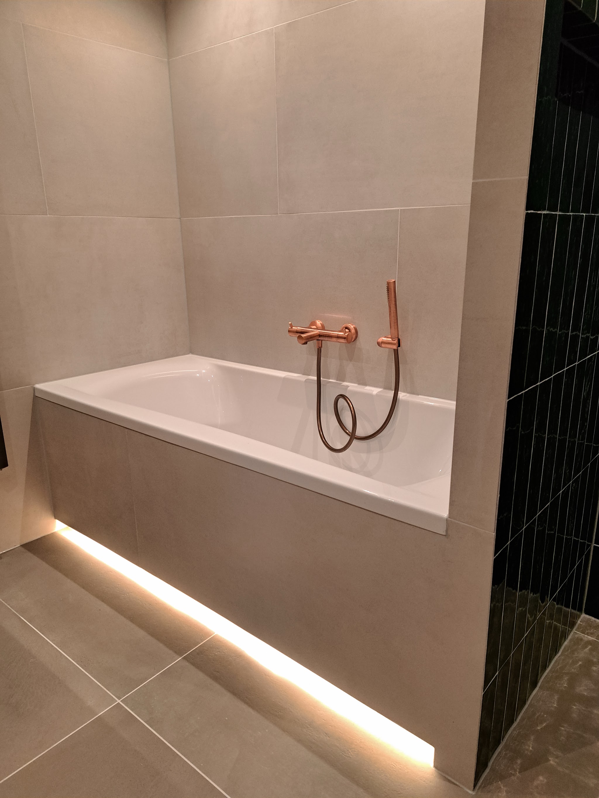 Badkamer met ligbad met verlichting onder de rand, koperen kranen en een groene tegelwand