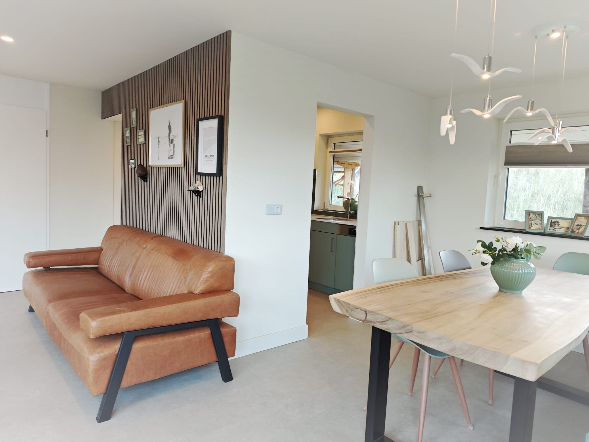Woonkamer met lamellenwand, leren bank en eettafel met zicht op een groene keuken